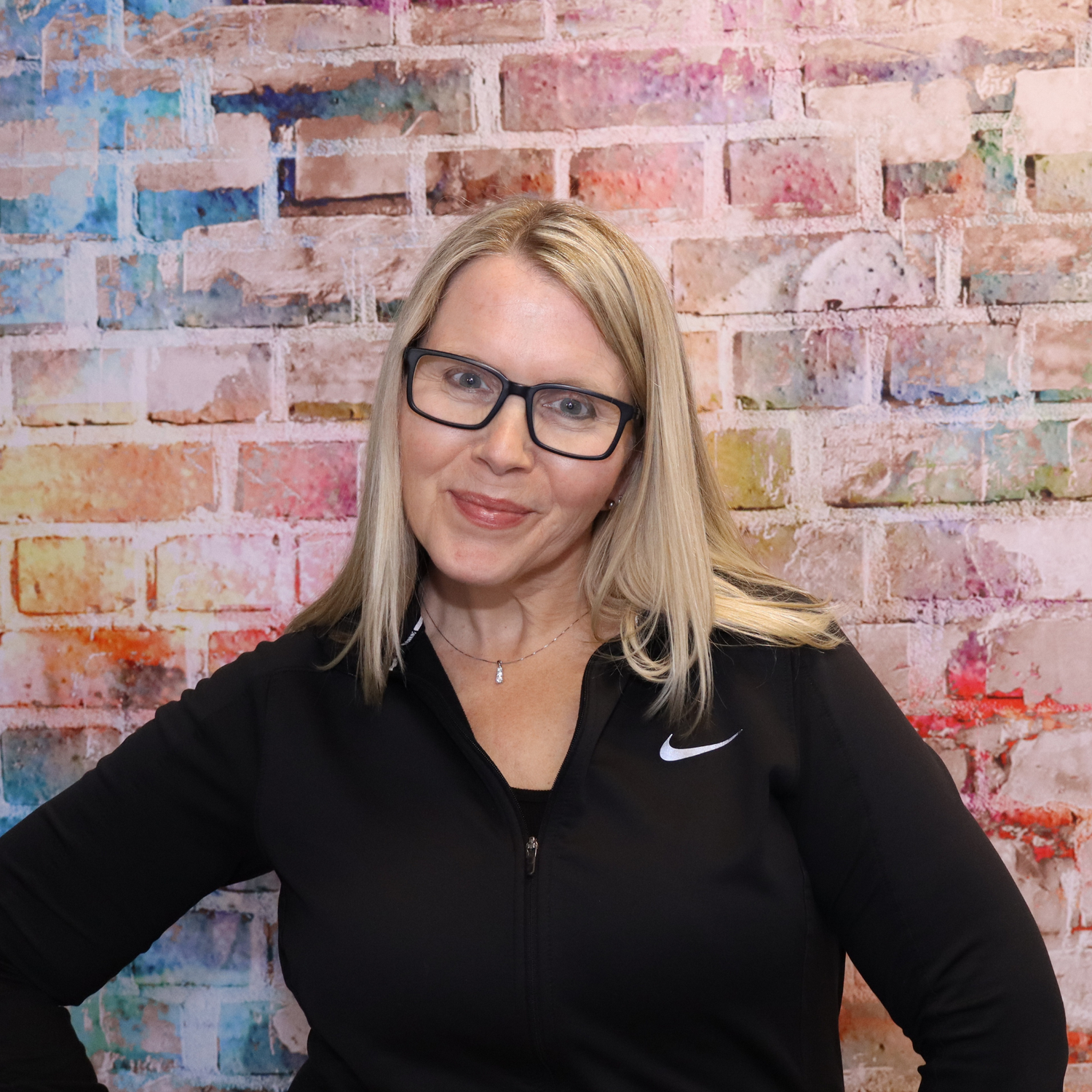 Woman wearing glasses and a black Nike jacket in front of a colorful brick wall